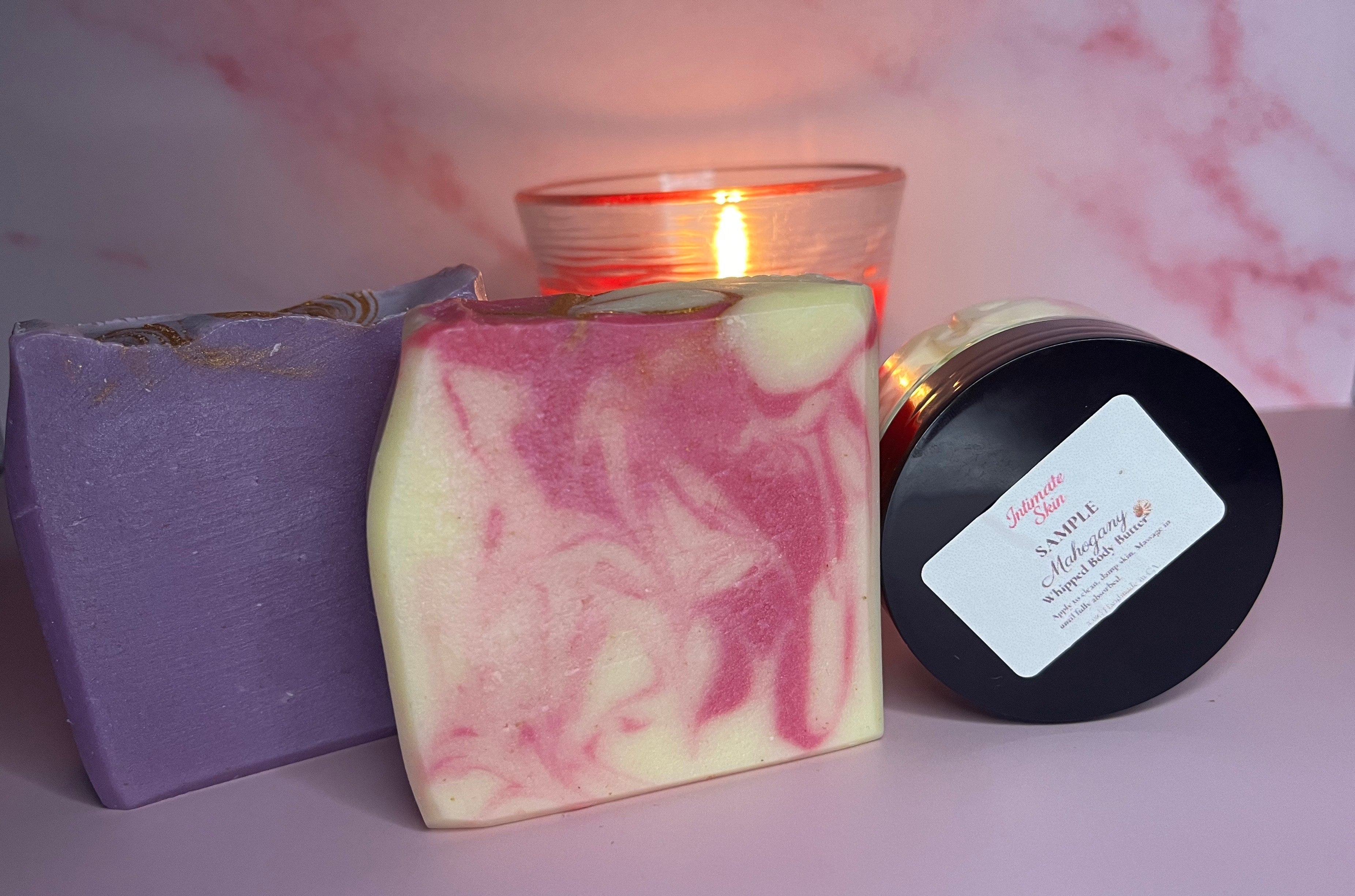Lavender and rose-colored soap bars displayed with a sample-sized body butter and a glowing candle on a blush pink background.
